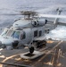 A U.S. Navy MH-60R Seahawk helicopter is hovering over the flight deck of a naval vessel at sea. The helicopter is armed with what appear to be AGM-114 Hellfire missiles on its starboard weapons pylon. The rotor blades are in motion, and the ocean is visible in the background with white wake from the ship.