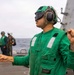 A naval flight deck crew member wearing green jersey and helmet with hearing protection is giving hand signals on what appears to be a military ship's flight deck. The green color of his jersey indicates he is likely a catapult and arresting gear officer or aircraft handling crew member. Additional personnel in various colored jerseys are visible in the background near the ship's superstructure.