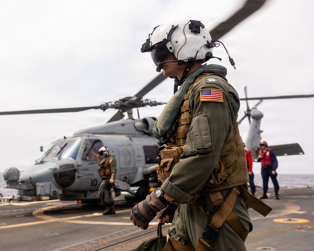 A U.S. military aviator in full flight gear, including a flight helmet with visor and tactical vest, walks across a ship's flight deck. In the background, a gray military helicopter (numbered '10') is visible with rotor blades spinning, along with additional crew members in various colored gear. The setting appears to be an active naval vessel operating at sea under overcast skies.
