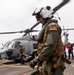 A U.S. military aviator in full flight gear, including a flight helmet with visor and tactical vest, walks across a ship's flight deck. In the background, a gray military helicopter (numbered '10') is visible with rotor blades spinning, along with additional crew members in various colored gear. The setting appears to be an active naval vessel operating at sea under overcast skies.