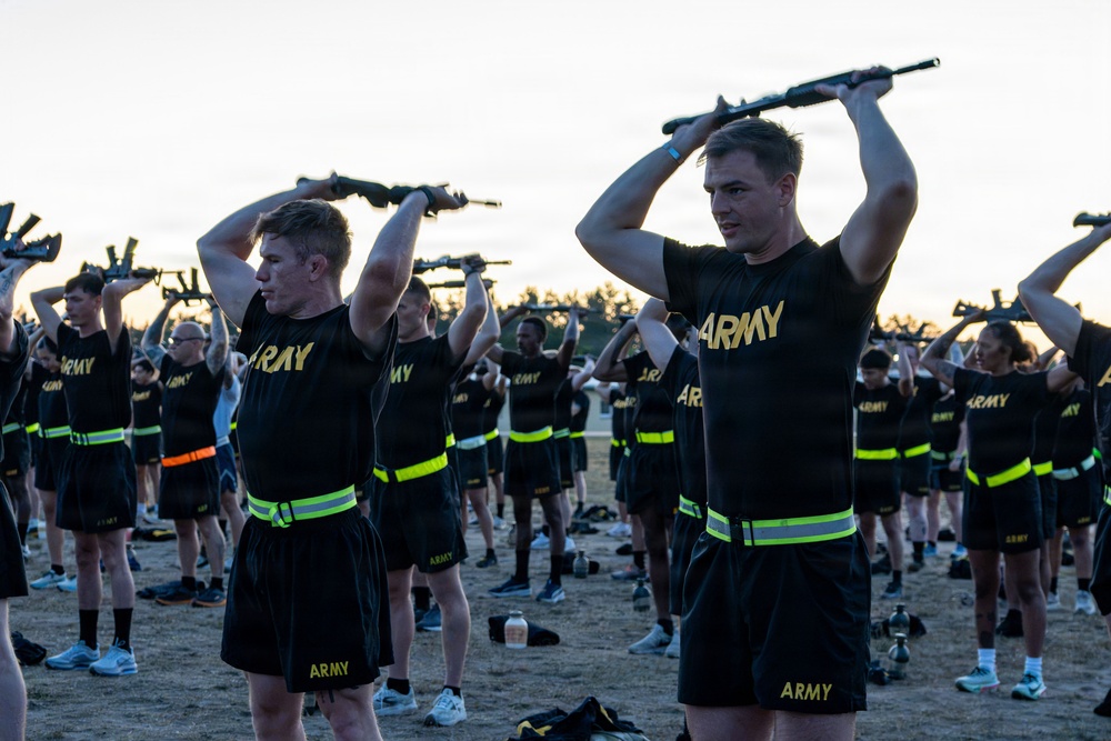 A large group of U.S. Army personnel are conducting a physical training exercise outdoors, holding rifles overhead in a synchronized drill. The soldiers are wearing standard Army PT (Physical Training) uniforms consisting of black shirts and shorts with gold 'ARMY' lettering and high-visibility yellow-green reflective belts. The scene appears to take place at dawn or dusk on an open field or parade ground.