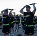 A large group of U.S. Army personnel are conducting a physical training exercise outdoors, holding rifles overhead in a synchronized drill. The soldiers are wearing standard Army PT (Physical Training) uniforms consisting of black shirts and shorts with gold 'ARMY' lettering and high-visibility yellow-green reflective belts. The scene appears to take place at dawn or dusk on an open field or parade ground.
