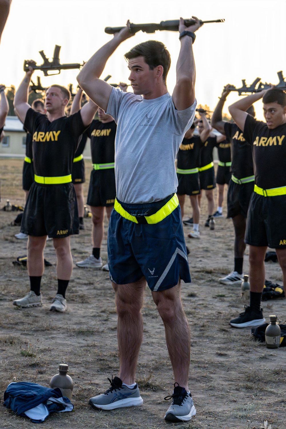 A group of military personnel are conducting physical training exercises outdoors, holding rifles overhead. The central figure is wearing U.S. Air Force PT gear (gray shirt and navy shorts with Air Force logo), while the surrounding personnel are wearing U.S. Army PT uniforms (black shirts with yellow 'ARMY' lettering). All participants are wearing high-visibility yellow safety belts around their waists.