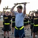 A group of military personnel are conducting physical training exercises outdoors, holding rifles overhead. The central figure is wearing U.S. Air Force PT gear (gray shirt and navy shorts with Air Force logo), while the surrounding personnel are wearing U.S. Army PT uniforms (black shirts with yellow 'ARMY' lettering). All participants are wearing high-visibility yellow safety belts around their waists.