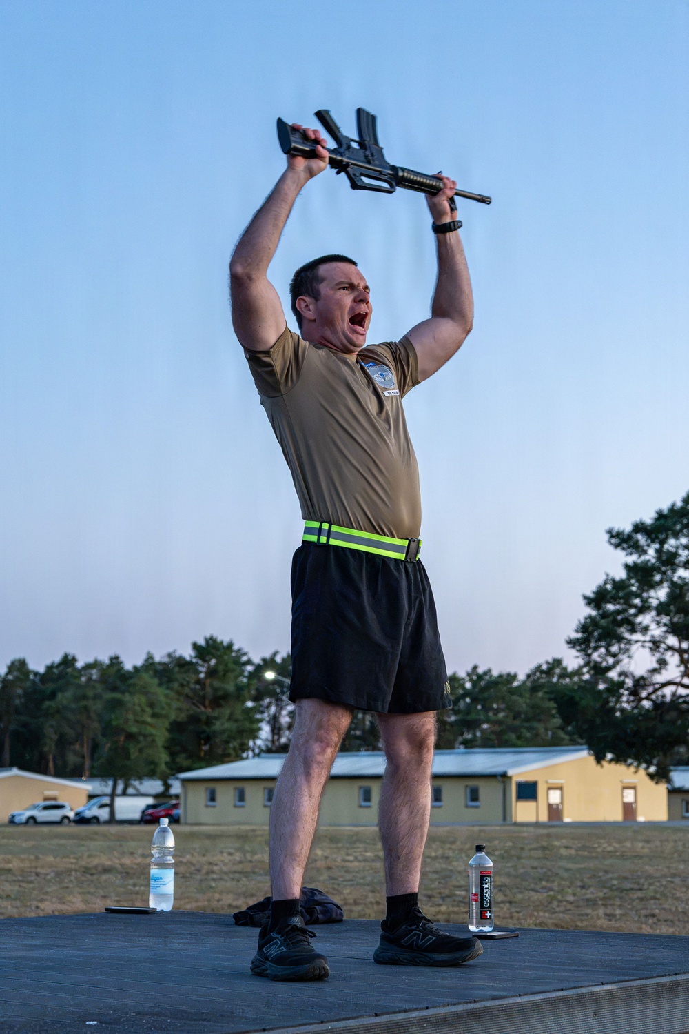 A man in military physical training attire stands on a platform outdoors, holding a rifle overhead with both hands while shouting. He is wearing an olive/tan PT shirt with a unit patch, black shorts, a high-visibility reflective belt, and New Balance athletic shoes. The setting appears to be a military installation based on the barracks-style buildings visible in the background.