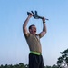 A man in military physical training attire stands on a platform outdoors, holding a rifle overhead with both hands while shouting. He is wearing an olive/tan PT shirt with a unit patch, black shorts, a high-visibility reflective belt, and New Balance athletic shoes. The setting appears to be a military installation based on the barracks-style buildings visible in the background.
