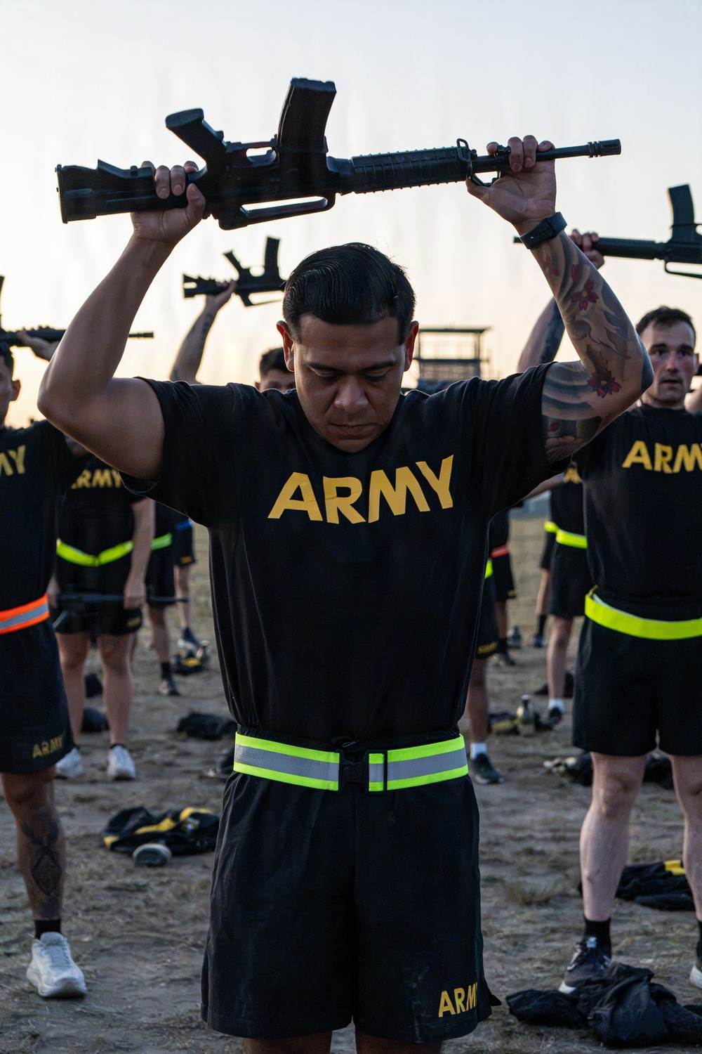 A group of U.S. Army soldiers in physical training (PT) uniforms are performing an exercise drill while holding rifles overhead. The foreground soldier, wearing a black Army PT shirt and shorts with a high-visibility reflective belt, holds what appears to be an M16/M4-style rifle above his head. Multiple soldiers in identical uniforms are visible in the background performing the same exercise on an outdoor dirt field.