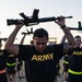 A group of U.S. Army soldiers in physical training (PT) uniforms are performing an exercise drill while holding rifles overhead. The foreground soldier, wearing a black Army PT shirt and shorts with a high-visibility reflective belt, holds what appears to be an M16/M4-style rifle above his head. Multiple soldiers in identical uniforms are visible in the background performing the same exercise on an outdoor dirt field.