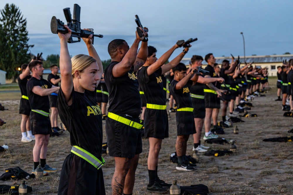 A large group of U.S. Army personnel in physical training uniforms are performing an exercise drill while holding rifles overhead with both arms extended. The activity appears to take place outdoors on a training field during early morning or dusk conditions. The participants are wearing matching black Army PT uniforms with yellow-green reflective belts.