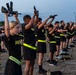 A large group of U.S. Army personnel in physical training uniforms are performing an exercise drill while holding rifles overhead with both arms extended. The activity appears to take place outdoors on a training field during early morning or dusk conditions. The participants are wearing matching black Army PT uniforms with yellow-green reflective belts.