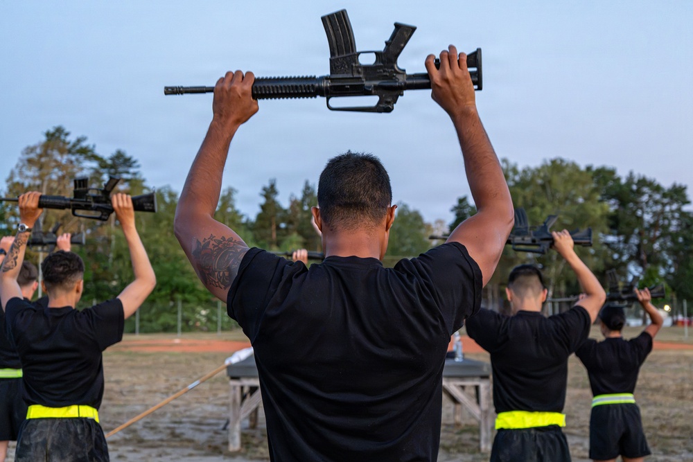 A group of military or law enforcement trainees are performing a physical training exercise outdoors, holding rifles overhead with both arms extended. The individuals are uniformly dressed in black t-shirts with yellow/neon reflective belts, suggesting a structured training environment. The setting appears to be a military base or training facility at dusk or early morning.