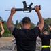 A group of military or law enforcement trainees are performing a physical training exercise outdoors, holding rifles overhead with both arms extended. The individuals are uniformly dressed in black t-shirts with yellow/neon reflective belts, suggesting a structured training environment. The setting appears to be a military base or training facility at dusk or early morning.