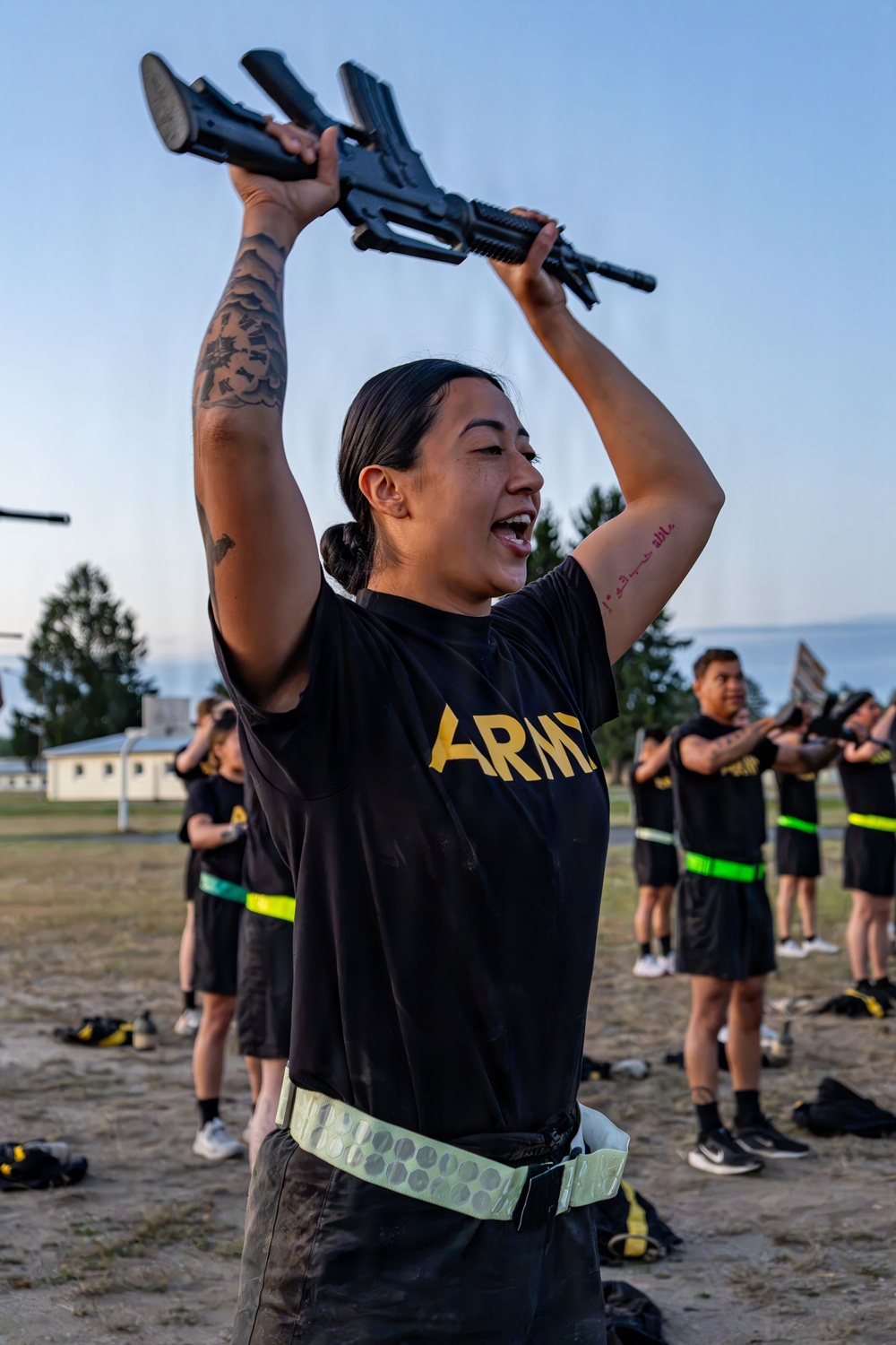 A female U.S. Army soldier in PT (physical training) gear holds a rifle overhead during what appears to be a drill or physical training exercise outdoors. She is smiling and appears to be calling out or responding to commands. Several other soldiers in matching black Army PT uniforms with reflective belts are visible in the background performing similar exercises.