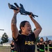 A female U.S. Army soldier in PT (physical training) gear holds a rifle overhead during what appears to be a drill or physical training exercise outdoors. She is smiling and appears to be calling out or responding to commands. Several other soldiers in matching black Army PT uniforms with reflective belts are visible in the background performing similar exercises.