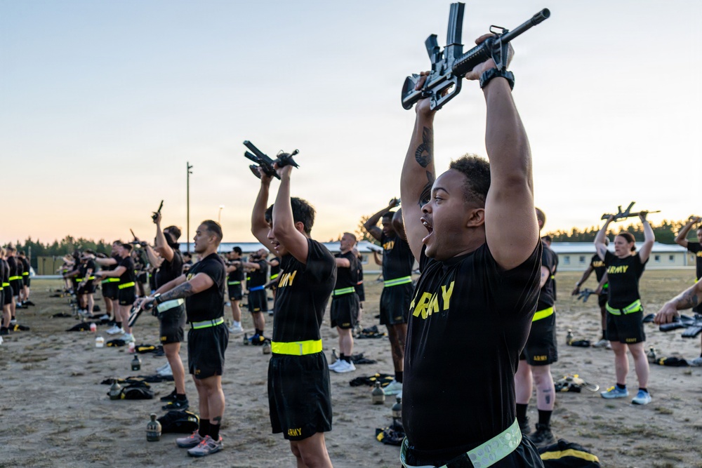 A large group of U.S. Army soldiers in physical training uniforms are performing an exercise drill on an outdoor sandy field, holding rifles overhead with both arms extended. The activity appears to be a structured physical fitness training session conducted at dawn or dusk, likely at a military installation. Soldiers are arranged in formation rows and are uniformly dressed in black Army PT gear with neon yellow-green accents.