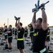 A large group of U.S. Army soldiers in physical training uniforms are performing an exercise drill on an outdoor sandy field, holding rifles overhead with both arms extended. The activity appears to be a structured physical fitness training session conducted at dawn or dusk, likely at a military installation. Soldiers are arranged in formation rows and are uniformly dressed in black Army PT gear with neon yellow-green accents.