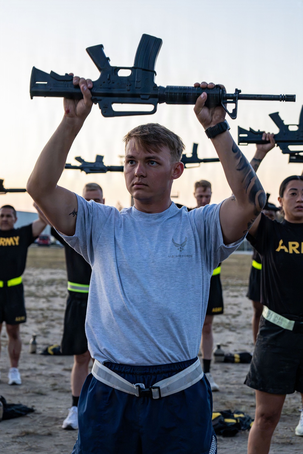 A group of military personnel are engaged in a physical training exercise, holding rifles overhead. The foreground subject wears a U.S. Air Force t-shirt while others in the background wear Army PT (physical training) uniforms. The setting appears to be an outdoor training area, likely during early morning hours based on the lighting.