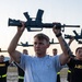A group of military personnel are engaged in a physical training exercise, holding rifles overhead. The foreground subject wears a U.S. Air Force t-shirt while others in the background wear Army PT (physical training) uniforms. The setting appears to be an outdoor training area, likely during early morning hours based on the lighting.