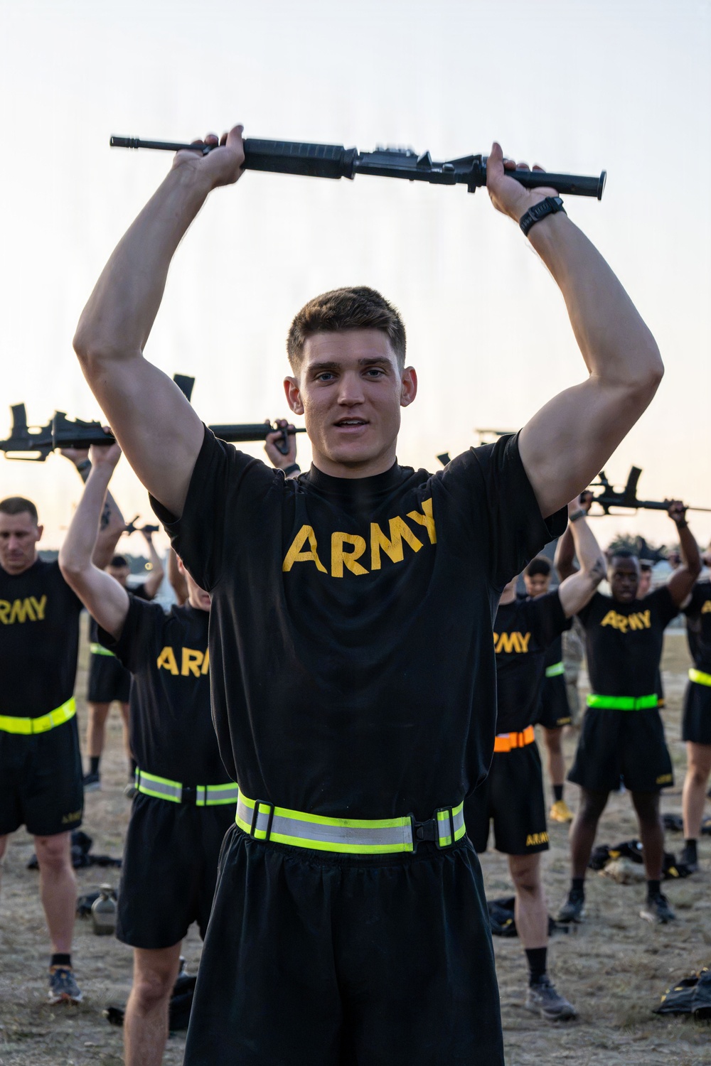 A group of U.S. Army soldiers in physical training (PT) uniforms are performing rifle exercises, holding what appear to be training rifles overhead. The foreground soldier, wearing a black 'ARMY' t-shirt with a reflective PT belt, is the primary focus of the image. Multiple soldiers in identical uniforms are visible in the background performing the same exercise.