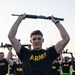 A group of U.S. Army soldiers in physical training (PT) uniforms are performing rifle exercises, holding what appear to be training rifles overhead. The foreground soldier, wearing a black 'ARMY' t-shirt with a reflective PT belt, is the primary focus of the image. Multiple soldiers in identical uniforms are visible in the background performing the same exercise.