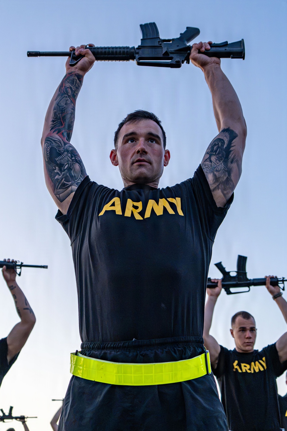 A U.S. Army soldier in physical training attire holds a rifle overhead with both arms extended, performing what appears to be a drill or PT exercise. The primary subject is heavily tattooed on both arms and wears a black Army PT shirt with yellow lettering and a high-visibility reflective belt. At least two other soldiers in identical uniforms are visible in the background performing the same exercise.
