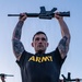 A U.S. Army soldier in physical training attire holds a rifle overhead with both arms extended, performing what appears to be a drill or PT exercise. The primary subject is heavily tattooed on both arms and wears a black Army PT shirt with yellow lettering and a high-visibility reflective belt. At least two other soldiers in identical uniforms are visible in the background performing the same exercise.