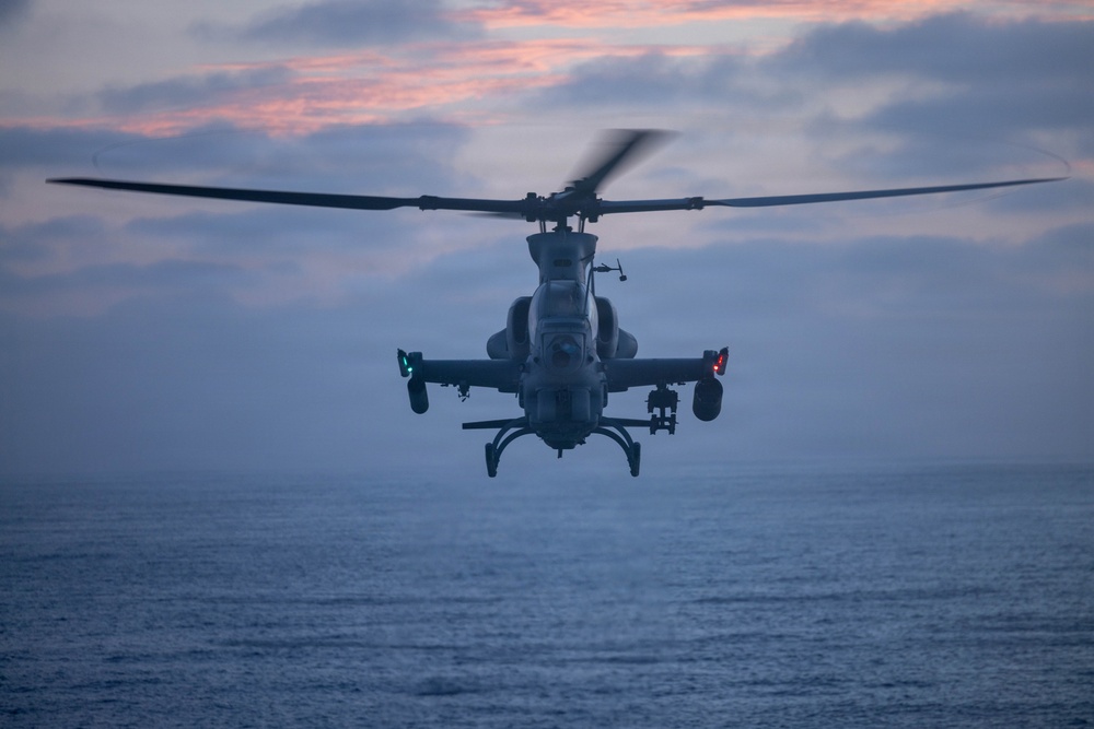 A military attack helicopter is photographed head-on in low-light conditions over open ocean water during dusk or dawn. The aircraft displays active navigation lights and appears to be in forward flight or hover. The silhouetted profile and tandem cockpit configuration are clearly visible against a partially cloudy twilight sky.