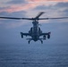 A military attack helicopter is photographed head-on in low-light conditions over open ocean water during dusk or dawn. The aircraft displays active navigation lights and appears to be in forward flight or hover. The silhouetted profile and tandem cockpit configuration are clearly visible against a partially cloudy twilight sky.