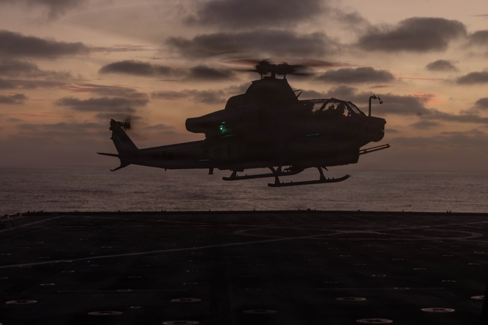 A military attack helicopter, consistent with an AH-1Z Viper or similar tandem-seat attack helicopter, is captured in silhouette hovering low over what appears to be a naval vessel's flight deck at dusk or dawn. The scene is set over open ocean with a dramatic twilight sky featuring orange and purple hues through cloud cover. A green navigation light is visible on the aircraft, and rotor blur indicates active flight.