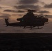 A military attack helicopter, consistent with an AH-1Z Viper or similar tandem-seat attack helicopter, is captured in silhouette hovering low over what appears to be a naval vessel's flight deck at dusk or dawn. The scene is set over open ocean with a dramatic twilight sky featuring orange and purple hues through cloud cover. A green navigation light is visible on the aircraft, and rotor blur indicates active flight.