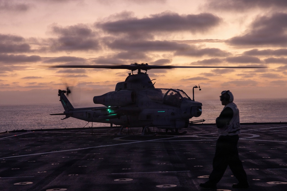 A military attack helicopter, identifiable as an AH-1Z Viper, sits on the flight deck of a naval vessel at dusk or dawn with rotors spinning. A single deck crew member in silhouette walks nearby, wearing hearing protection. The scene is dramatically lit by a sunset/sunrise sky over open ocean.