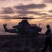 A military attack helicopter, identifiable as an AH-1Z Viper, sits on the flight deck of a naval vessel at dusk or dawn with rotors spinning. A single deck crew member in silhouette walks nearby, wearing hearing protection. The scene is dramatically lit by a sunset/sunrise sky over open ocean.