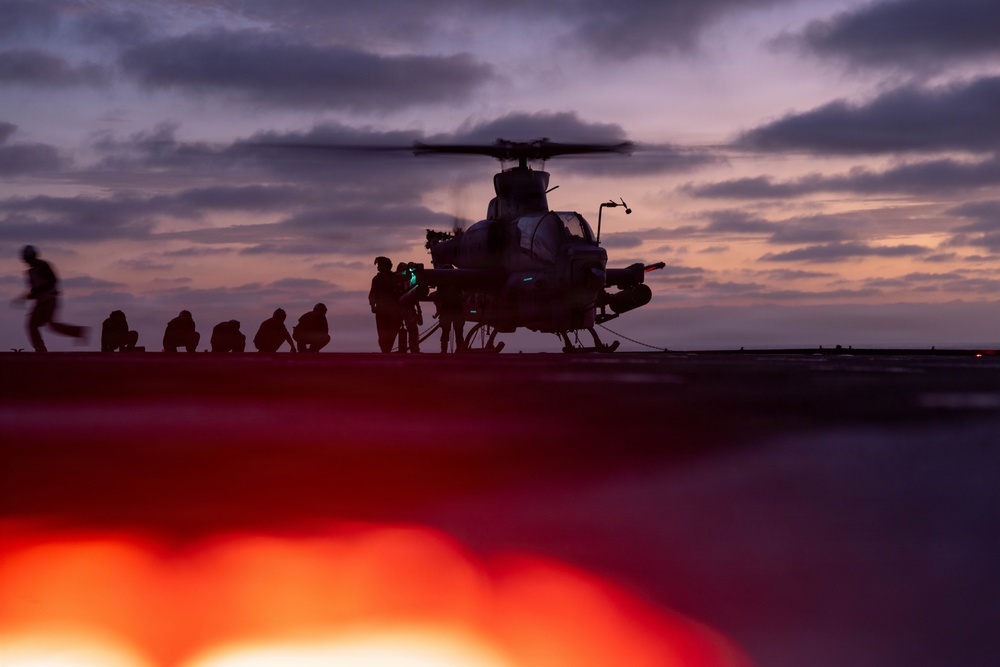 A military helicopter, appearing to be an attack helicopter (likely an AH-1Z Viper based on silhouette), is on the deck of a naval vessel at dusk or dawn. Ground crew personnel are visible as silhouettes around the aircraft, suggesting pre-flight or post-flight operations. The scene is dramatically lit by a purple-orange twilight sky and a bright red light source in the foreground.