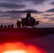 A military helicopter, appearing to be an attack helicopter (likely an AH-1Z Viper based on silhouette), is on the deck of a naval vessel at dusk or dawn. Ground crew personnel are visible as silhouettes around the aircraft, suggesting pre-flight or post-flight operations. The scene is dramatically lit by a purple-orange twilight sky and a bright red light source in the foreground.