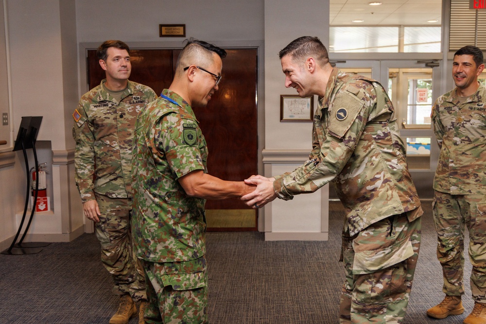 The image shows a military greeting ceremony taking place indoors, where a soldier in Japanese Ground Self-Defense Force (JGSDF) camouflage uniform is shaking hands with a U.S. Army soldier in OCP (Operational Camouflage Pattern) uniform. Two additional U.S. Army soldiers are visible in the background observing the exchange. The setting appears to be a military building lobby or reception area.