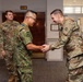The image shows a military greeting ceremony taking place indoors, where a soldier in Japanese Ground Self-Defense Force (JGSDF) camouflage uniform is shaking hands with a U.S. Army soldier in OCP (Operational Camouflage Pattern) uniform. Two additional U.S. Army soldiers are visible in the background observing the exchange. The setting appears to be a military building lobby or reception area.