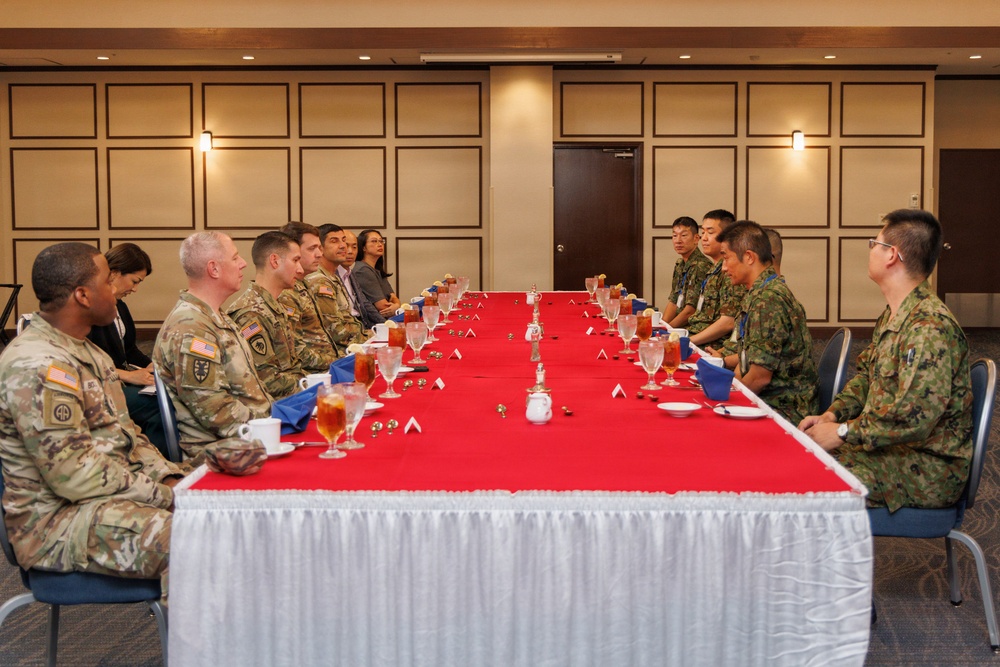 A formal military meeting or luncheon is taking place at a long rectangular table covered with a red tablecloth and white skirting. Personnel in U.S. Army camouflage uniforms (OCP pattern) are seated on the left side, facing personnel in what appears to be Japanese Ground Self-Defense Force camouflage uniforms on the right side. The setting suggests a bilateral military exchange or liaison meeting in a conference or banquet room.