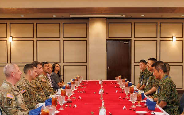 A formal military meeting or luncheon is taking place at a long rectangular table covered with a red tablecloth and white skirting. Personnel in U.S. Army camouflage uniforms (OCP pattern) are seated on the left side, facing personnel in what appears to be Japanese Ground Self-Defense Force camouflage uniforms on the right side. The setting suggests a bilateral military exchange or liaison meeting in a conference or banquet room.