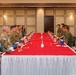 A formal military meeting or luncheon is taking place at a long rectangular table covered with a red tablecloth and white skirting. Personnel in U.S. Army camouflage uniforms (OCP pattern) are seated on the left side, facing personnel in what appears to be Japanese Ground Self-Defense Force camouflage uniforms on the right side. The setting suggests a bilateral military exchange or liaison meeting in a conference or banquet room.
