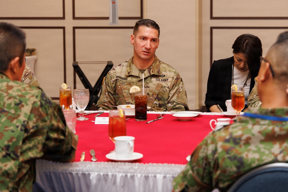 A U.S. Army soldier identified by name tape as 'HUGGINS' is seated at a formal dining table with red tablecloth, engaged in conversation with personnel wearing what appear to be Japanese Ground Self-Defense Force camouflage uniforms. A civilian woman in a black jacket is seated nearby, appearing to take notes. The setting suggests a formal military liaison or bilateral meeting meal.
