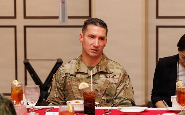 A U.S. Army soldier identified by name tape as 'HUGGINS' is seated at a formal dining table with red tablecloth, engaged in conversation with personnel wearing what appear to be Japanese Ground Self-Defense Force camouflage uniforms. A civilian woman in a black jacket is seated nearby, appearing to take notes. The setting suggests a formal military liaison or bilateral meeting meal.