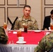 A U.S. Army soldier identified by name tape as 'HUGGINS' is seated at a formal dining table with red tablecloth, engaged in conversation with personnel wearing what appear to be Japanese Ground Self-Defense Force camouflage uniforms. A civilian woman in a black jacket is seated nearby, appearing to take notes. The setting suggests a formal military liaison or bilateral meeting meal.