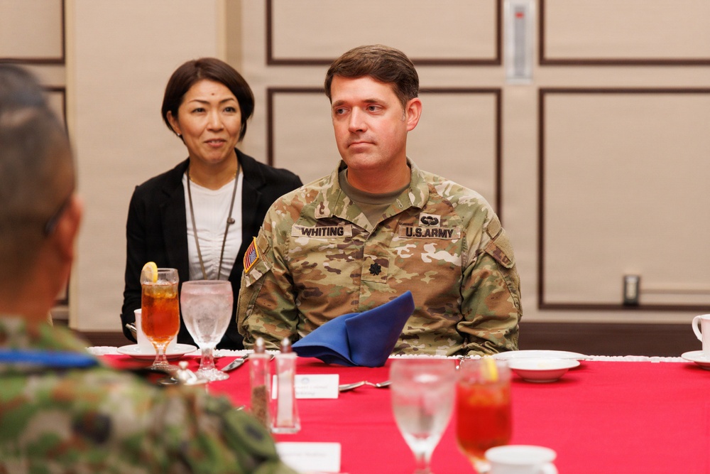 A U.S. Army soldier identified by name tape as 'WHITING' is seated at a formal dining table alongside a civilian woman wearing a lanyard/badge. The setting appears to be an official military dining event or meeting, likely involving multinational participants given the visible camouflage uniform of another individual across the table. The table is set with a red tablecloth, glassware, and place settings consistent with a formal luncheon or dinner engagement.