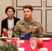 A U.S. Army soldier identified by name tape as 'WHITING' is seated at a formal dining table alongside a civilian woman wearing a lanyard/badge. The setting appears to be an official military dining event or meeting, likely involving multinational participants given the visible camouflage uniform of another individual across the table. The table is set with a red tablecloth, glassware, and place settings consistent with a formal luncheon or dinner engagement.