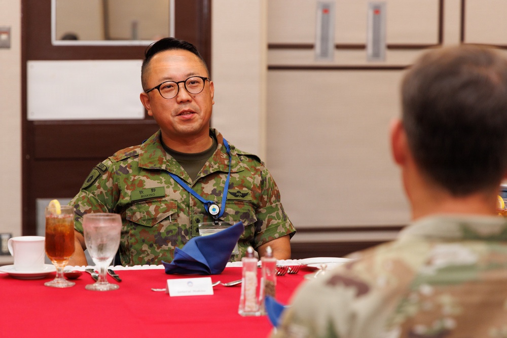 Two military personnel are seated at a formally set dining table with a red tablecloth, engaged in conversation. The individual facing the camera wears a Japanese Ground Self-Defense Force (JGSDF) camouflage uniform with a name tag reading 'MAKINO' and a blue lanyard with a badge. The person with their back to the camera appears to wear a US military MultiCam/OCP uniform, suggesting a bilateral military meeting or luncheon.