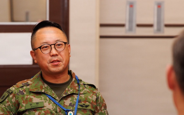 Two military personnel are seated at a formally set dining table with a red tablecloth, engaged in conversation. The individual facing the camera wears a Japanese Ground Self-Defense Force (JGSDF) camouflage uniform with a name tag reading 'MAKINO' and a blue lanyard with a badge. The person with their back to the camera appears to wear a US military MultiCam/OCP uniform, suggesting a bilateral military meeting or luncheon.