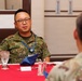 Two military personnel are seated at a formally set dining table with a red tablecloth, engaged in conversation. The individual facing the camera wears a Japanese Ground Self-Defense Force (JGSDF) camouflage uniform with a name tag reading 'MAKINO' and a blue lanyard with a badge. The person with their back to the camera appears to wear a US military MultiCam/OCP uniform, suggesting a bilateral military meeting or luncheon.
