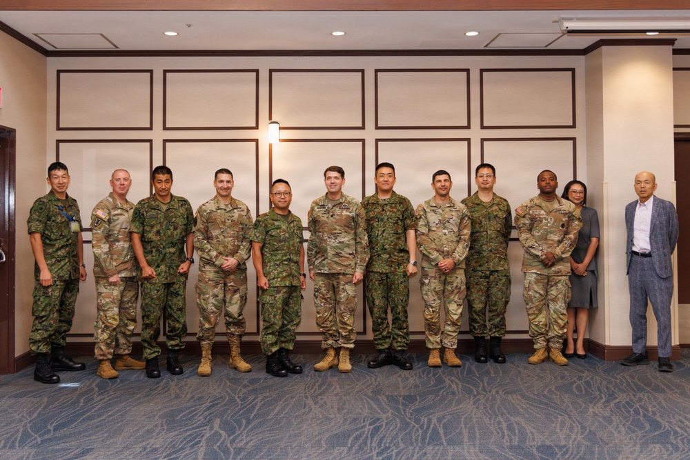A formal group photograph of approximately 13 individuals, the majority wearing military uniforms of two distinct camouflage patterns, suggesting a multinational military gathering or bilateral meeting. Two civilians, one woman and one man in business attire, stand at the far right of the group. The setting appears to be an indoor conference or hotel facility with decorative wall paneling.