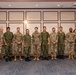 A formal group photograph of approximately 13 individuals, the majority wearing military uniforms of two distinct camouflage patterns, suggesting a multinational military gathering or bilateral meeting. Two civilians, one woman and one man in business attire, stand at the far right of the group. The setting appears to be an indoor conference or hotel facility with decorative wall paneling.