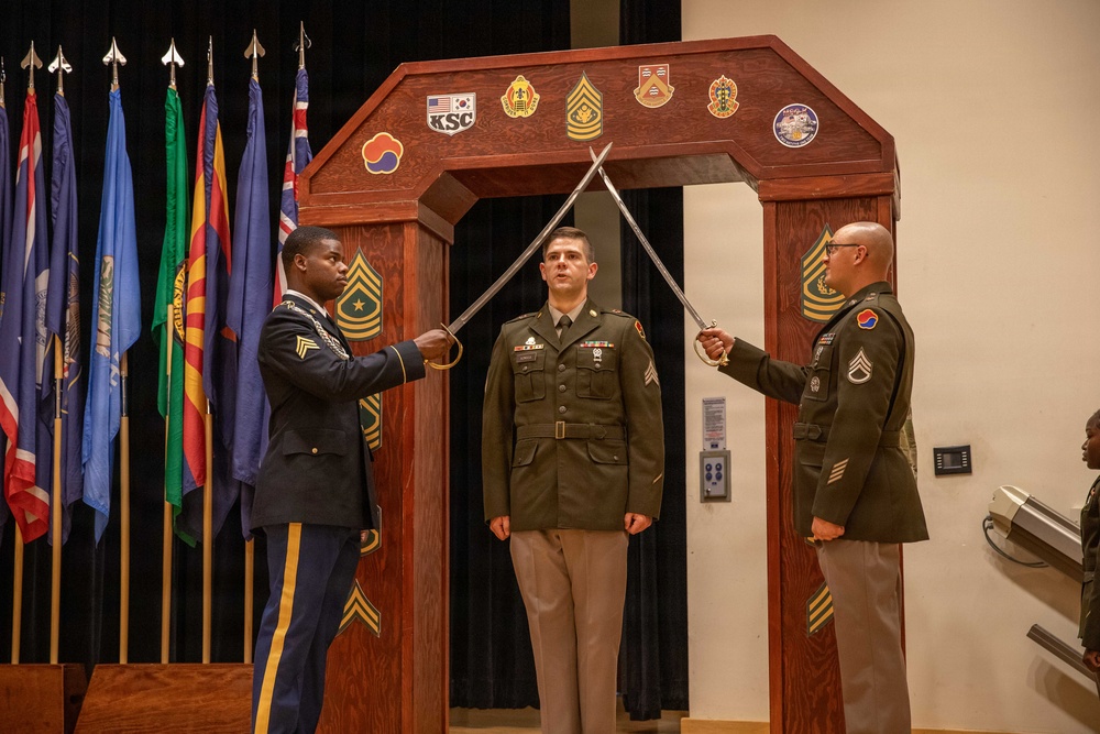 A U.S. Army military ceremony is taking place, likely a promotion or induction ceremony. Two soldiers in dress uniforms are crossing swords over a third soldier standing beneath a ceremonial wooden arch decorated with unit patches and insignia. Multiple military flags are displayed in the background.